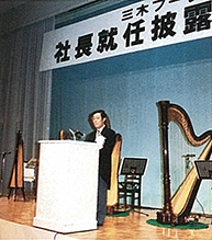 三木プーリの沿革写真 新社長就任お披露目会の様子（演台で挨拶を行う新社長と会場ステージの風景）